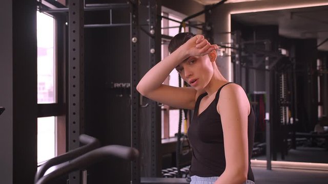 Closeup shoot of young attractive athlete female with ponytail working out on the orbitrek sweating and being tired looking at camera in the gym indoors