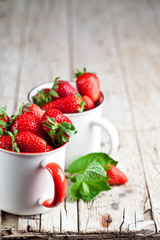 Organic red strawberries in two white ceramic cups and mint leaves on rustic wooden background. Healthy sweet food, vitamins and fruity concept.