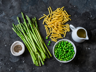 Ingredients for cooking vegetarian lunch - raw pasta, asparagus, green peas on a dark background, top view