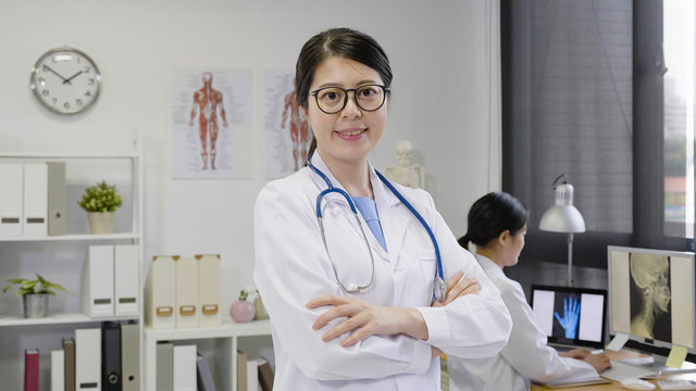 confident medical woman doctor at hospital face camera smiling attractive. happy female nurse standing crossed arms looking right. coworker in uniform concentrated working on computer in background.