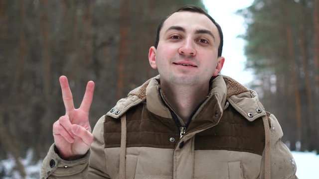 Portrait of handsome man in jacket is showing victory gesture at camera. Man stands in winter forest