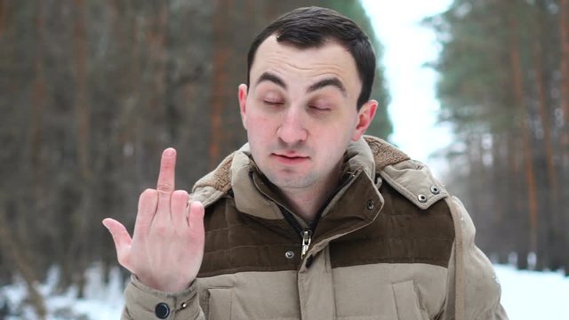 Portrait of young man in jacket is showing middle finger. Man stands in winter forest