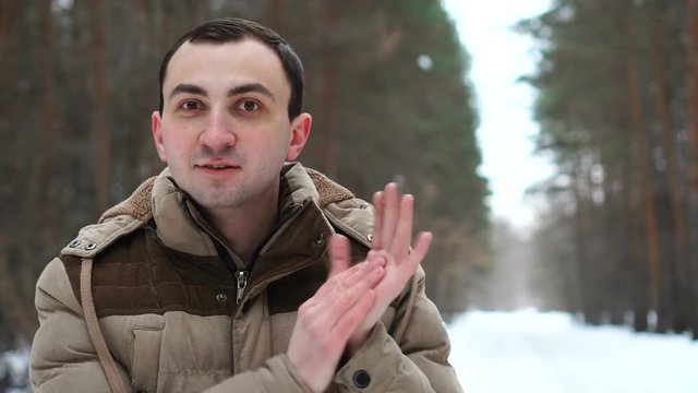 Portrait of young man in jacket is applauding outdoors. Man stands in winter forest