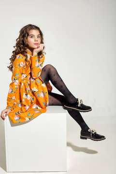Fashion for girls 10 years in a yellow dress posing in the Studio on a cube. Curly hair, black shoes.