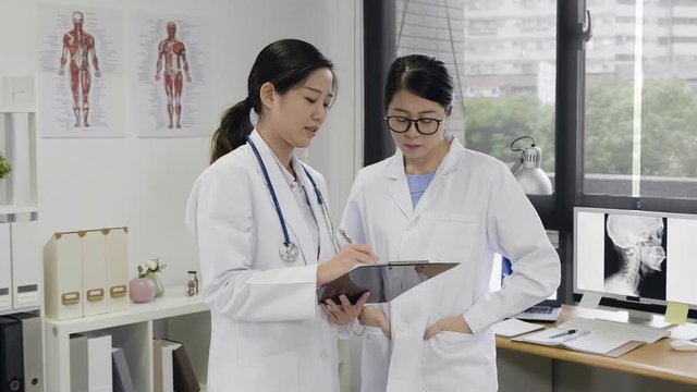 two women doctors at hospital with clipboard standing near window in modern office discussing. young girls medical staff workers in uniform working concentrated consult in clinic ub afternoon.