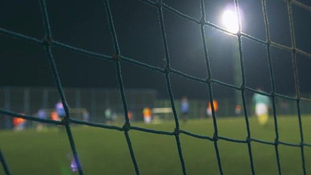 Evening football game. Football players play on the training field, illuminated by stadium lights. View through goal net. 