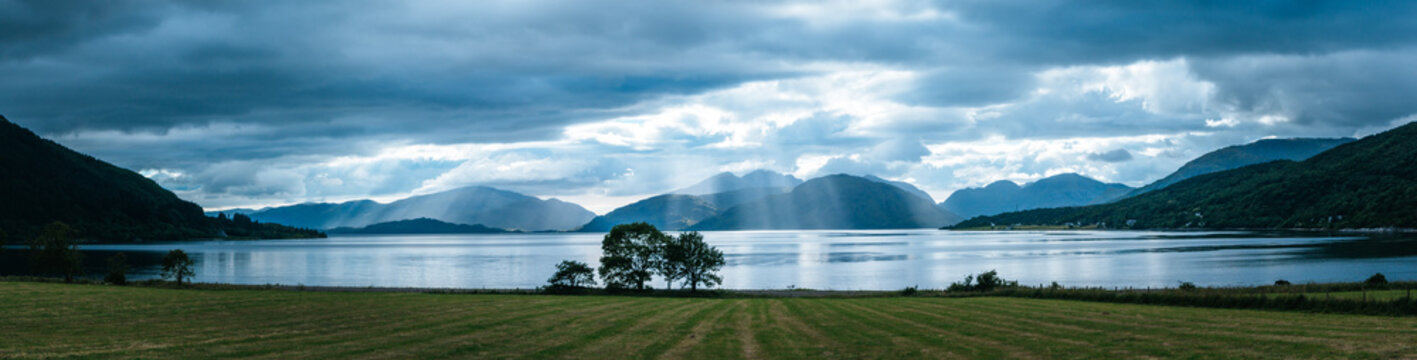Mystic landscape lake scenery in Scotland: Cloudy sky, meadow, trees and lake with sunbeams, mountain range in the background. Loch Linnhe.
