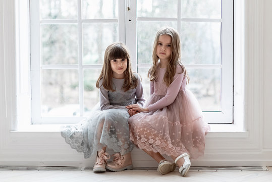Two little girls with big eyes sit on a windowsill near a large window