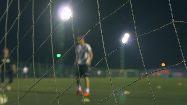 Evening football game. Football players play on the training field, illuminated by stadium lights. View through goal net. 