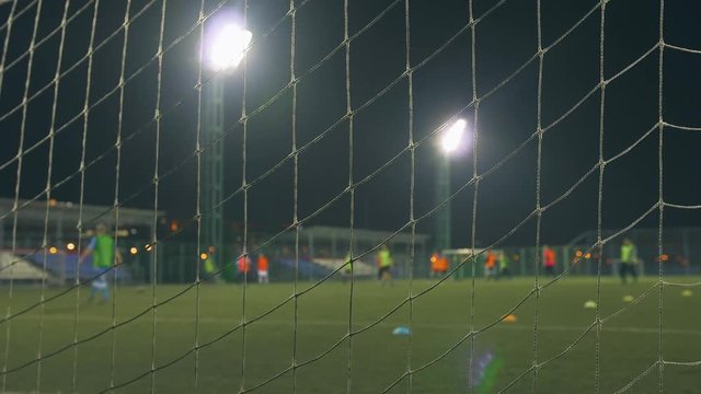 Evening football game. Football players play on the training field, illuminated by stadium lights. View through goal net. 