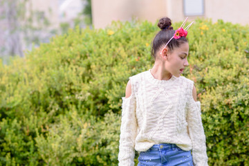 Portrait beautiful young teenager wearing flower crown, white sweater and stylish jeans is standing and looking at the side in the city park near the green trees. Charming sweet girl. Mixed race model