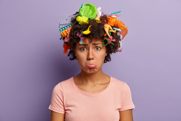 Troubled dissatisfied lady purses lower lip, collects plastic garbage, wears casual t shirt, being eco friendly, upset by serious environmental problem, has trash in curly hair isolated on purple wall