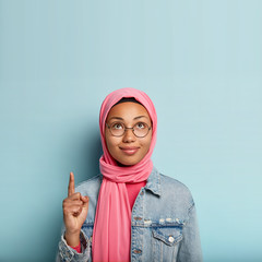 Charming young woman with dark healthy skin, looks and points up, indicates with index finger, wears pink hijab, fashionable jacket, stands over blue wall, advertises copy space, shows right way