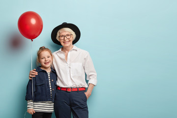 Family portrait of dranddaughter and granny embrace and celebrate holiday, hold air ballonon, wear festive clothes, express positive emotions isolated on blue studio wall. Generation and fest concept