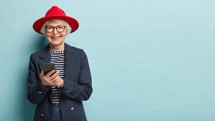 Overjoyed senior lady in stylish apparel and hat chats with friends online, happy to learn using...