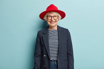 Trendy fashionable elderly woman smiles happily, shows white teeth, has wrinkled skin, dressed in...