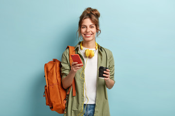 Studio shot of glad female student has coffee break after lectures, listens audio book in headphones, enjoys record from website, uses mobile phone for chatting online, has rucksack on back. - obrazy, fototapety, plakaty
