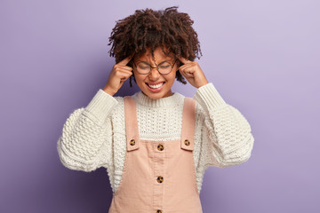 Nervous dark skinned lady manages stress, relieves pain, keeps fore fingers on temples, smirks...
