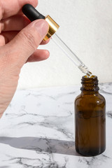 Pipette dropper with drop of natural oil above brown glass bottle on marble background. Natural organic skin care beauty products.