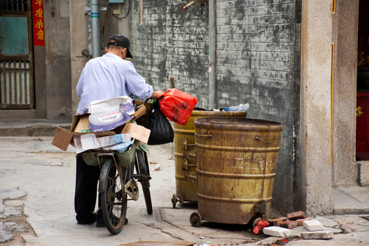 Chinese old man trash keeper find and pickup recyclable waste in garbage bin to basket of bicycle in small alley at Shantou or Swatow city in Guangdong, China