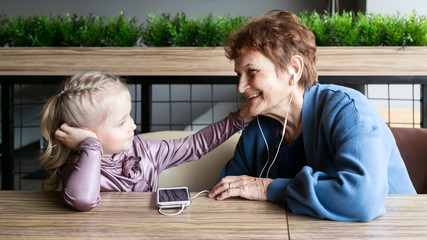An elderly retired woman on a smartphone turned on an audio tale  little girl in headphones. Grandmother wore headphones to her granddaughter to listen to the audio tale.