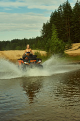 Beautiful blonde girl on a orang ATV rides on the river, standing on the mountain. Around the forest, sand and blue sky.She rides on a green swamp, in the mud and all wet. She's wearing a blue bikini.