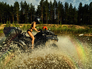 Beautiful blonde girl on a black ATV rides on the river, standing on the mountain. Around the forest, sand and blue sky.She rides on a green swamp, in the mud and all wet. She's wearing a blue bikini.