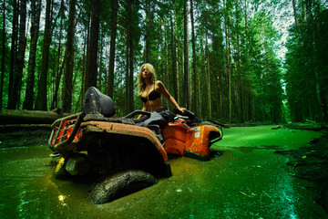 Piękna blondynki dziewczyna na orang ATV jedzie na rzece, stoi na górze. Wokół lasu, piasku i niebieskiego nieba, jeździ na zielonym bagnie, w błocie i cała mokra. Ona ma na sobie niebieskie bikini. - 264766375