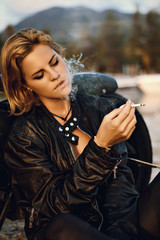 Plakat Young woman biker smoking seat next to motorcycle on the road right side and sunset, mountain background, nobody