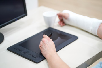 Artist with broken arm drawing something with graphic tablet. Hold pen in broken fractured arm in plaster cast.
