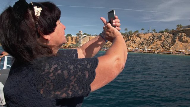Senior female happy adult tourist on a boat uses a mobile smart phone to get photos of a tropical island. Slow Motion.