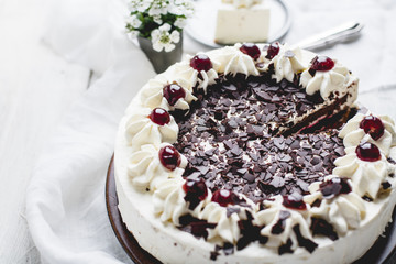 German, delicious Black Forest cake, with a delicate white cream, cherries in alcohol and dark chocolate on a white wooden background decorated with white linen fabric and white flowers