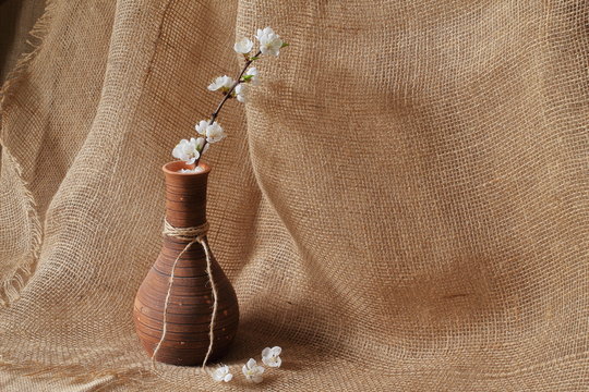 Branch of apple tree with white beautiful flowers in original clay vase on rough homespun jute background. Concept is style, elegance and beauty.