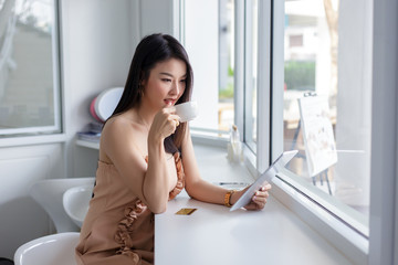 Beautiful asian woman who enjoy playing tablet and drinking coffee in a white coffee shop.