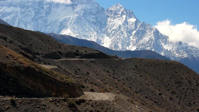 A 4x4 vehicle travels along a distant mountain road with the snow capped Upper Mustang of the Himalayas rising in the background.