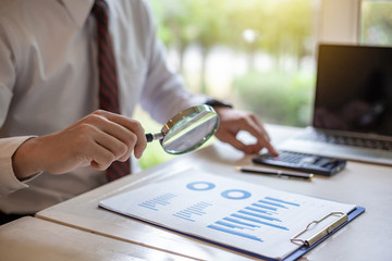 Businessman holding a magnifying glass with graph information document on the desk.