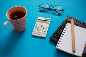 Top view of blue office desk, with mug of tea, pencil and empty notebook on keyboard, glasses and white calculator.