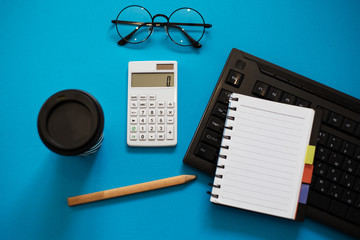Top view of blue office desk, with cup of coffee, pencil and empty notebook on keyboard, glasses and white calculator.