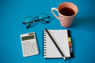 Top view of blue office desk, with cup of tea, empty notebook and pencil, glasses and white calculator.