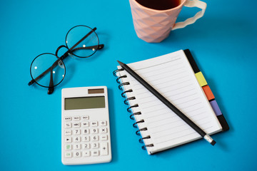 Top view of blue office desk, with cup of tea, empty notebook and pencil, glasses and white calculator.