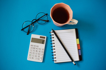 Top view of blue office desk, with cup of tea, empty notebook and pencil, glasses and white calculator.