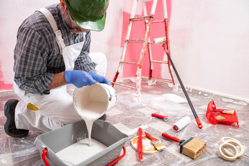 Caucasian house painter worker in white overalls, with helmet and goggles pour the white paint into...
