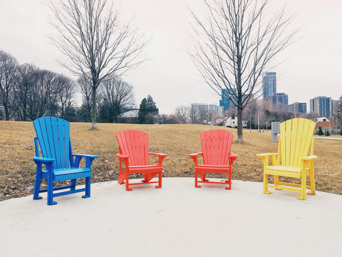 Four Colorful Red, Blue And Yellow Wooden Muskoka Adirondack Chairs Standing In Row In Park Outside On Spring Autumn Day. Concept Of Relaxation And Calm Talk Conversation Among Friends Or Family.