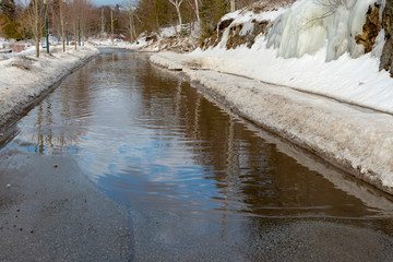 A flooded road in winter. Snowbanks are beside the road, and the sky is reflected in the water that covers the road.