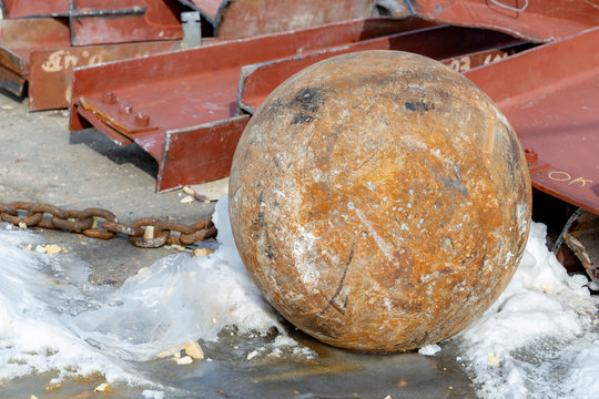 A rusty wrecking ball lying on the ground. A small amount of snow is around it, and steel beams are in the background.