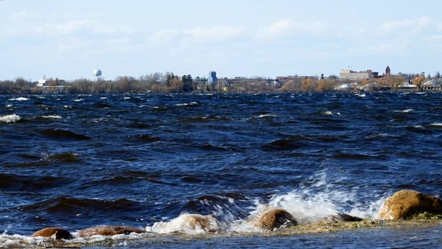 Bemidji, Minnesota is seen across Lake Irving as foaming waves roll onto shoreline over boulders on a windy afternoon.