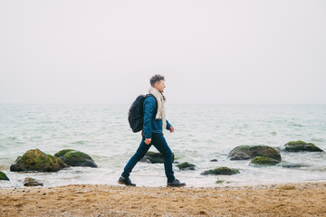 Traveler with a backpack standing near a rock against a beautiful sea with waves, a stylish hipster boy posing near a calm ocean during a wonderful journey around the world. Vertical