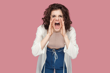 Portrait of beautiful brunette young woman with curly hairstyle in casual style standing, looking at camera and screaming. people expression and emotion. indoor studio shot isolated on pink background