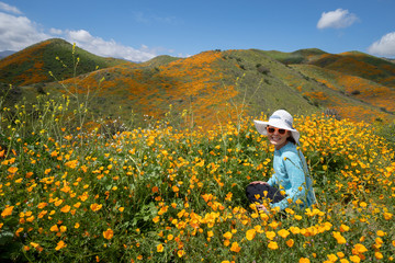 Fototapeta premium Young woman wearing sunglasses and straw hat poses in poppy field in California