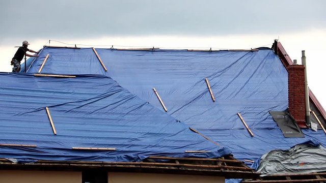 The roofer works on roof when is rain. The tarp covers the roof of the old house in the reconstruction.